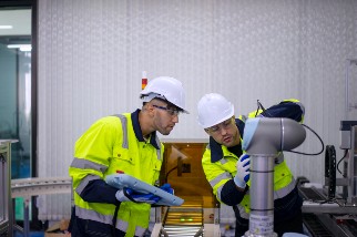 Two individuals in safety gear are working with a robotic arm in an industrial setting. They are wearing high-visibility jackets and helmets, focusing on operating or adjusting the machinery.