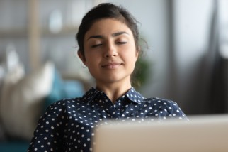 Young woman safety professional who is stressed working from home is taking a break to meditate in front of her laptop