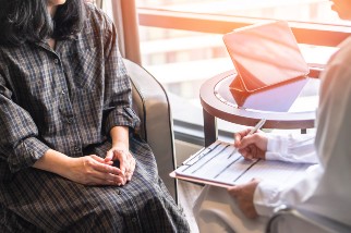 Two individuals are seated near a window in an office setting; one is holding a clipboard and taking notes while the other, dressed in a plaid shirt, is seated facing them.