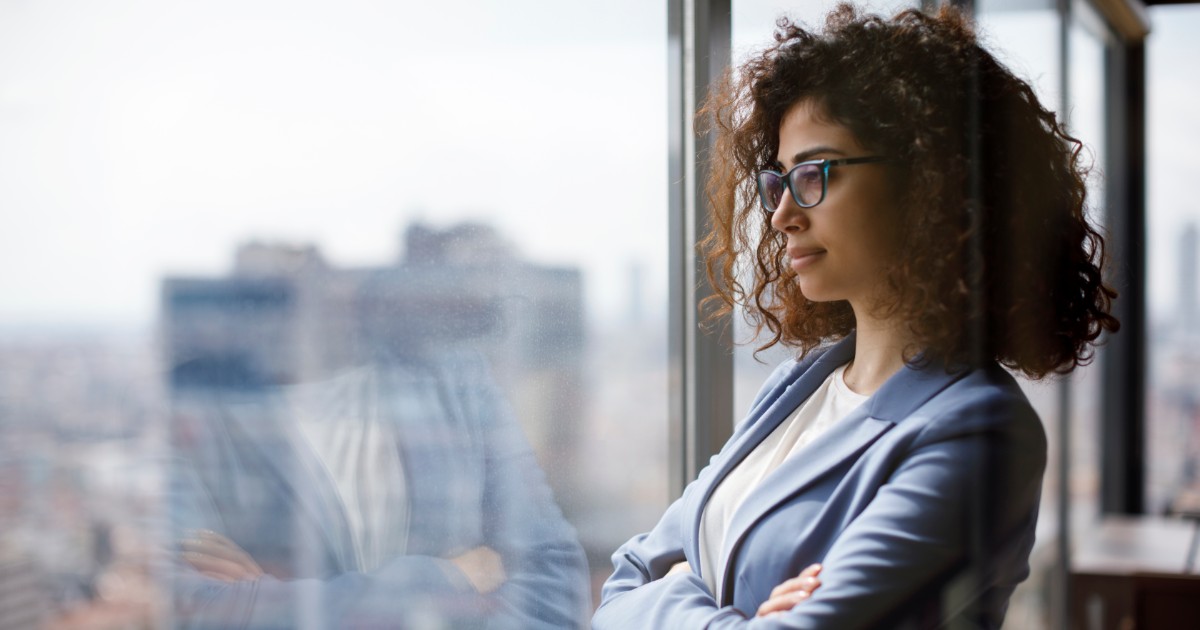 Young safety professional woman with glasses standing in an office alone and looking out the window