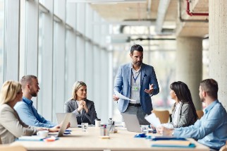 A person stands at the head of a conference table, speaking to five seated individuals in a modern office with floor-to-ceiling windows. Laptops and documents are visible on the table.