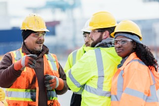 Four construction workers in safety gear, including bright orange and yellow vests and helmets, are having a conversation at an outdoor site. The background shows industrial equipment.