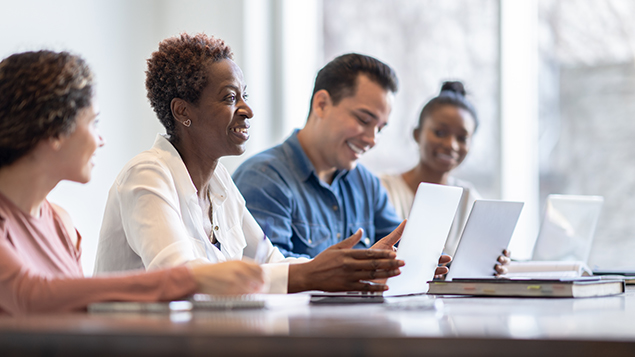 A group of adults are sitting at a classroom table with laptops