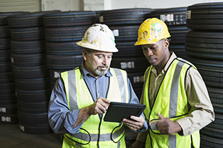 Two multi-ethnic men in a warehouse with stacks of truck tires, using a digital tablet. They are wearing yellow safety vests and hard hats, standing in front of the tires. The older man, in his 50s, is holding the tablet, pointing to the screen.